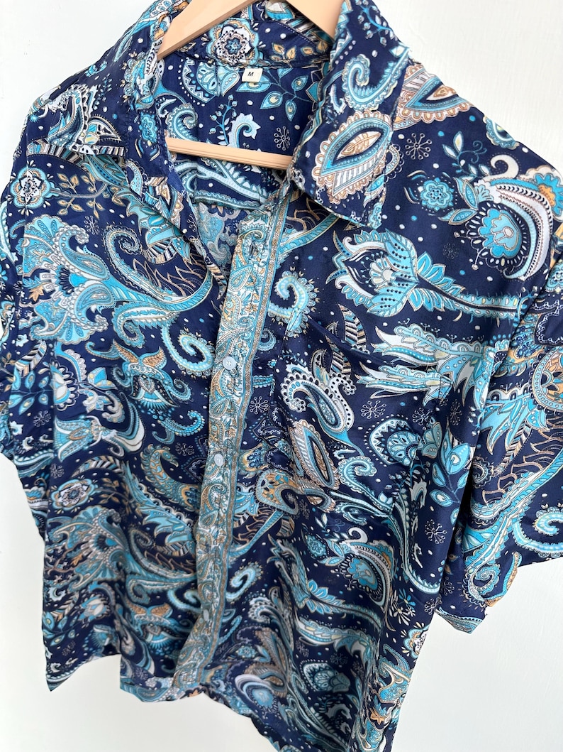 Op de afbeelding: Een overhemd met korte mouwen met een paisley patroon in marineblauw, lichtblauw en bruin. Het overhemd hangt aan een houten hanger. Het heeft een kraag en een knoopsluiting. Maat M.