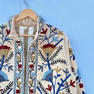 Pu&ograve; includere: Una giacca di velluto color crema con un ricamo floreale in tonalit&agrave; di blu, rosso e oro. La giacca ha un colletto alto e una chiusura con cerniera. L'etichetta della taglia indica "XL".