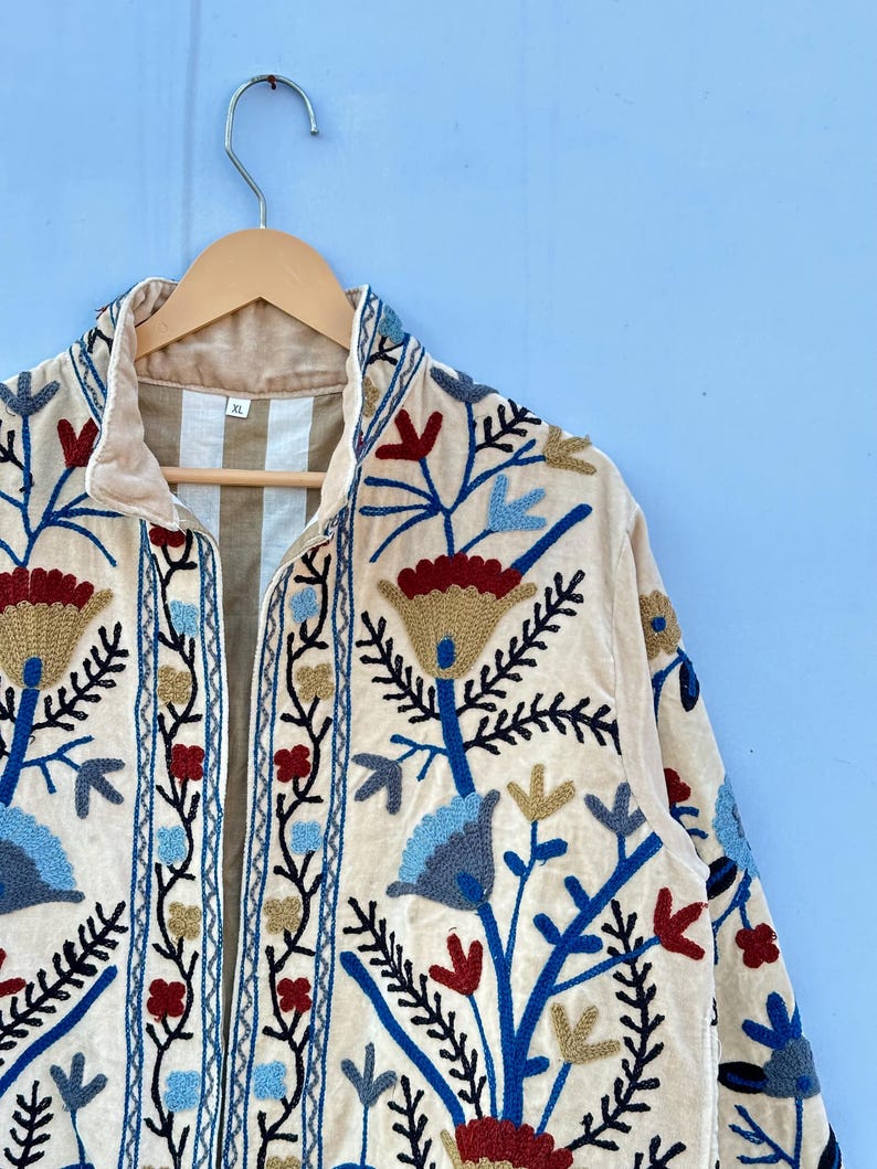 Pu&ograve; includere: Una giacca di velluto color crema con un ricamo floreale in tonalit&agrave; di blu, rosso e oro. La giacca ha un colletto alto e una chiusura con cerniera. L'etichetta della taglia indica "XL".
