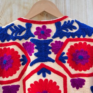 May include: Close-up of a cream-coloured embroidered jacket on a wooden hanger. The jacket features vibrant red, blue, and purple floral and geometric patterns. The embroidery is detailed and the colours are bold.