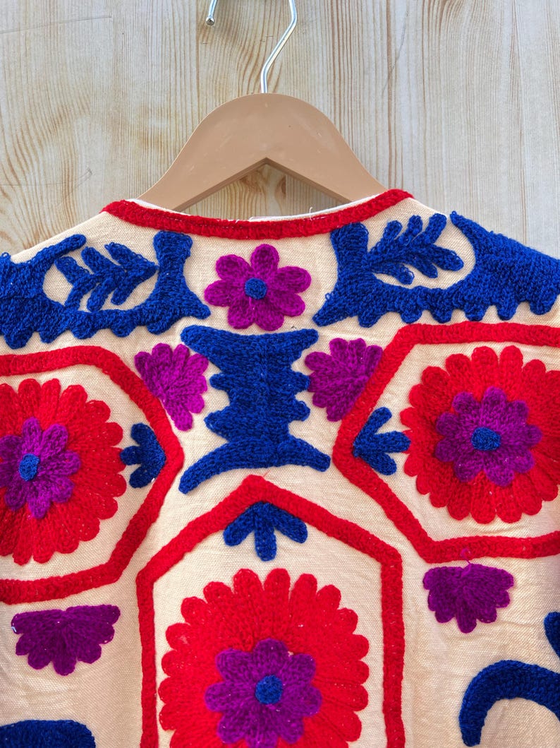 May include: Close-up of a cream-coloured embroidered jacket on a wooden hanger. The jacket features vibrant red, blue, and purple floral and geometric patterns. The embroidery is detailed and the colours are bold.