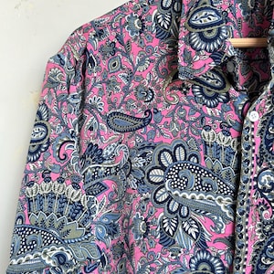 Op de afbeelding: Een roze paisley overhemd met knopen. Het overhemd heeft een gedetailleerd ontwerp in blauw, grijs en wit. Het overhemd met lange mouwen hangt aan een houten kleerhanger.