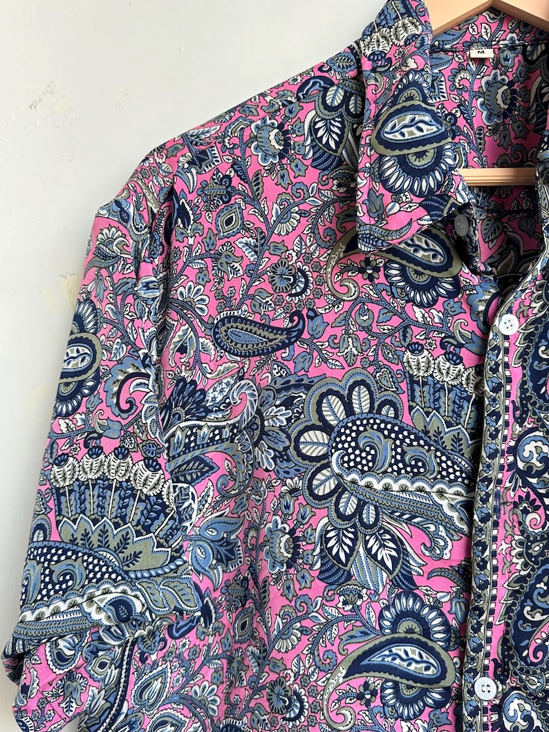 Op de afbeelding: Een roze paisley overhemd met knopen. Het overhemd heeft een gedetailleerd ontwerp in blauw, grijs en wit. Het overhemd met lange mouwen hangt aan een houten kleerhanger.