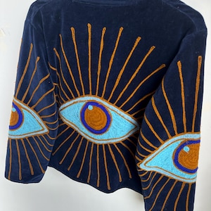 May include: Navy blue velvet top with a high collar. The top features embroidered eye designs in light blue, orange, and brown. The eye designs are on the sleeves and back of the top.