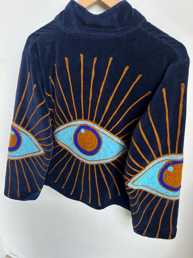 May include: Navy blue velvet top with a high collar. The top features embroidered eye designs in light blue, orange, and brown. The eye designs are on the sleeves and back of the top.
