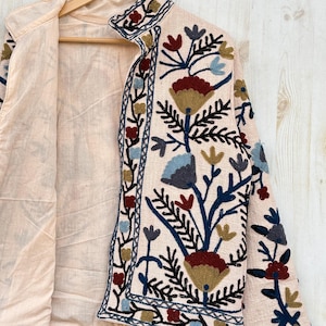 Pode incluir: Casaco bege claro com bordados florais intrincados em tons de vermelho, azul e dourado. O casaco apresenta uma gola alta e &eacute; forrado com um tecido bege mais claro. O design floral est&aacute; concentrado na frente e nas mangas.