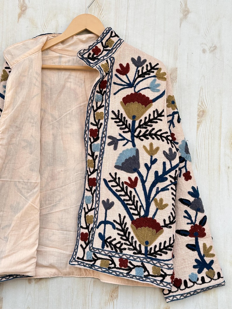 Pode incluir: Casaco bege claro com bordados florais intrincados em tons de vermelho, azul e dourado. O casaco apresenta uma gola alta e &eacute; forrado com um tecido bege mais claro. O design floral est&aacute; concentrado na frente e nas mangas.