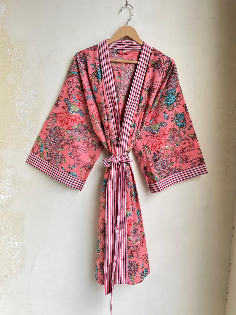Pu&ograve; includere: Accappatoio rosa con stampa floreale e cravatta abbinata, appeso a una gruccia di legno. L'accappatoio presenta un motivo ripetuto di fiori e foglie nelle tonalit&agrave; del rosa, del verde acqua e del rosso. I bordi e la cravatta hanno strisce rosa e bianche.