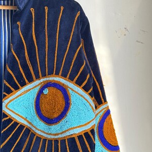 May include: Navy blue velvet jacket with embroidered eye design. The jacket features a large eye motif in shades of turquoise, brown, and blue, with gold-colored sunburst rays emanating from the eye. The interior lining has a blue and white striped pattern.