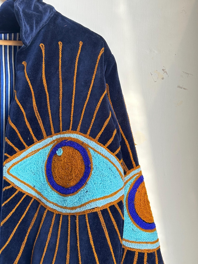 May include: Navy blue velvet jacket with embroidered eye design. The jacket features a large eye motif in shades of turquoise, brown, and blue, with gold-colored sunburst rays emanating from the eye. The interior lining has a blue and white striped pattern.