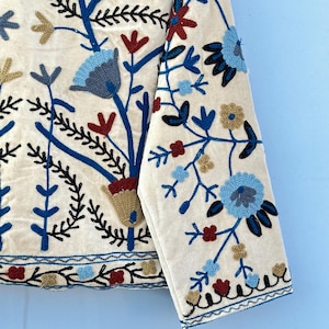 Pu&ograve; includere: Una giacca di cotone bianca con ricami floreali blu, rossi e dorati. La giacca ha maniche lunghe e chiusura con bottoni.