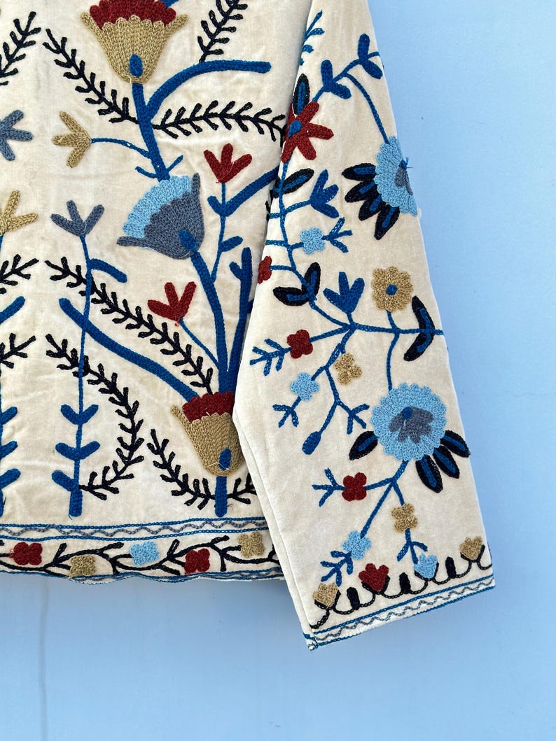 Pu&ograve; includere: Una giacca di cotone bianca con ricami floreali blu, rossi e dorati. La giacca ha maniche lunghe e chiusura con bottoni.