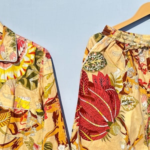 Op de afbeelding: Een beige pyjamaset met een bloemenprint met rode, gele en groene bloemen. Het bovenstuk heeft een knoopsluiting en een kraag. De broek heeft een elastische tailleband.