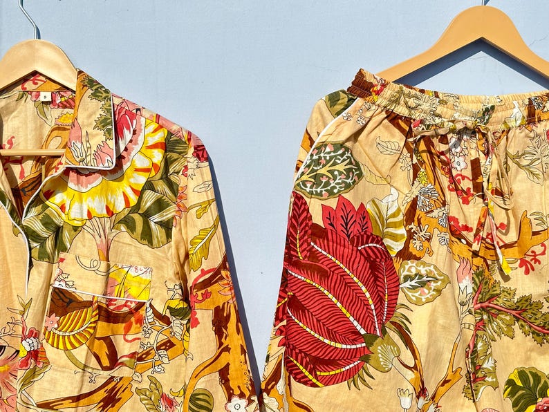 Op de afbeelding: Een beige pyjamaset met een bloemenprint met rode, gele en groene bloemen. Het bovenstuk heeft een knoopsluiting en een kraag. De broek heeft een elastische tailleband.