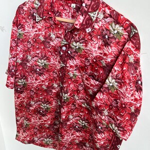 Op de afbeelding: Een shirt met korte mouwen en een opvallend bloemenpatroon in rood, bordeauxrood en wit. Het shirt hangt aan een houten hanger, wat de all-over bloemenprint benadrukt. Het patroon loopt door over de kraag en manchetten.