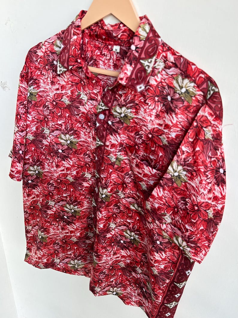 Op de afbeelding: Een shirt met korte mouwen en een opvallend bloemenpatroon in rood, bordeauxrood en wit. Het shirt hangt aan een houten hanger, wat de all-over bloemenprint benadrukt. Het patroon loopt door over de kraag en manchetten.