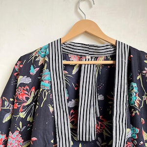 Pu&ograve; includere: Accappatoio kimono con stampa floreale nera con fiori e farfalle rossi, turchese e rosa. L'accappatoio ha un bordo a righe in bianco e nero. L'accappatoio &egrave; appeso a una gruccia di legno.