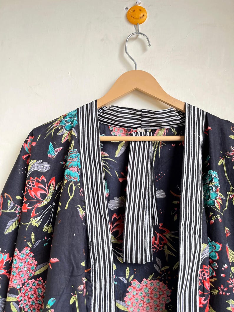 Pu&ograve; includere: Accappatoio kimono con stampa floreale nera con fiori e farfalle rossi, turchese e rosa. L'accappatoio ha un bordo a righe in bianco e nero. L'accappatoio &egrave; appeso a una gruccia di legno.