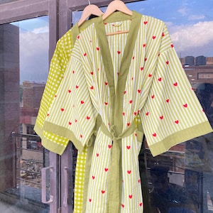 May include: Two lightweight robes hanging on wooden hangers. One robe is lime green with a white gingham pattern. The other robe is white with lime green stripes and red hearts. Both robes have a matching belt and trim.