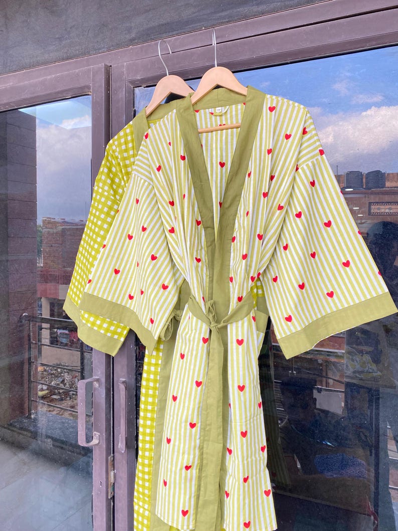 May include: Two lightweight robes hanging on wooden hangers. One robe is lime green with a white gingham pattern. The other robe is white with lime green stripes and red hearts. Both robes have a matching belt and trim.