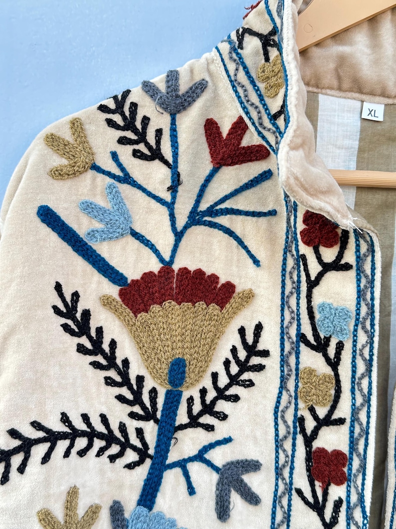 Pu&ograve; includere: Una giacca di velluto color crema con un ricamo floreale in tonalit&agrave; di blu, rosso, verde e marrone. La giacca ha un colletto e una chiusura con bottoni. L'etichetta della taglia sulla giacca riporta "XL".