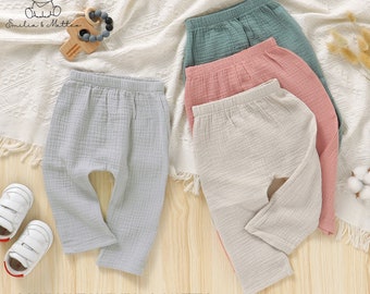 May include: Four pairs of baby pants in pastel colors: light gray, pink, beige, and teal. The pants are made of a soft, textured fabric. The image also includes a pair of white baby shoes, a wooden toy, and the text "COPYRIGHT BY EMILIA & MATTEO".