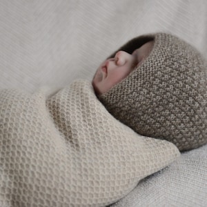 Könnte beinhalten: Ein Baby trägt eine braune Strickmütze mit Wabenmuster, eingewickelt in eine beige Strickdecke mit Wabenmuster.