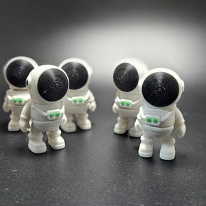 May include: Five white astronaut figurines with black visors and green details. The figurines are standing on a grey surface.