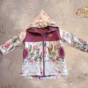 Könnte beinhalten: Eine rosa-weiße Kapuzenjacke mit Blumendruck und Reißverschluss. Die Jacke hat zwei Taschen vorne und eine Kapuze mit rosa Futter.