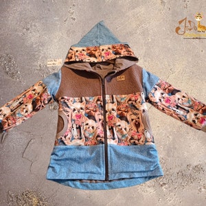 Könnte beinhalten: Eine braune und blaue Kapuzenjacke mit Reißverschluss. Die Jacke hat einen bunten Druck mit Cartoon-Hunden auf den Ärmeln und der Vorderseite. Die Kapuze ist mit einem blauen Stoff gefüttert.