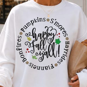 Op de afbeelding: Een wit sweatshirt met lange mouwen en een ronde hals, voorzien van een cirkelvormige afbeelding met de tekst "happy Fall y'all!" in zwarte schrijfletters. De omringende tekst bevat "Pumpkins", "S'mores", "Hayrides", "Flannels" en "Bonfires" met kleine bladpictogrammen.