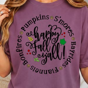 Op de afbeelding: Een pruimkleurig t-shirt met een cirkelvormige afbeelding en de zwarte tekst "happy fall y'all!" in het midden. Rondom de tekst staan de woorden "pumpkins", "S'mores", "Hayrides", "Flannels" en "Bonfires" met kleine blad- en pooticoontjes.