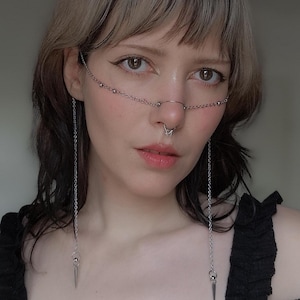 Puede incluir: Un accesorio facial de cadena plateada con cadenas colgantes y colgantes de púas. La cadena se extiende por la nariz y la frente, con pequeñas cuentas y un piercing en la nariz. La modelo lleva un top negro.