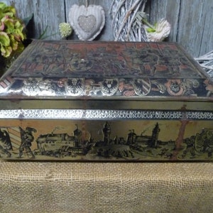 Könnte beinhalten: Eine rechteckige antike Metallbox mit Scharnierdeckel. Die Box zeigt detaillierte Illustrationen von Figuren, Gebäuden und Wappen in Schwarz und Gold. Die Box hat einen dekorativen Rand und eine rotbraune Oberseite.