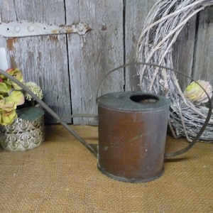 Könnte beinhalten: Eine antike Gießkanne aus Kupfer mit einem langen, gebogenen Ausguss und einem Griff. Die Kanne ist rostig und hat eine Patina des Alters. Sie steht auf einer Jutestoff-Oberfläche vor einer Holztür und einem Kranz.
