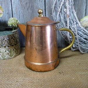 Könnte beinhalten: Eine Kupferkanne mit einem Messinggriff und -deckel. Die Kanne steht auf einer Jutestoff-Oberfläche.