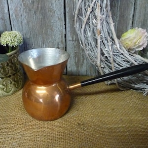 Könnte beinhalten: Eine Kaffeekanne aus Kupfer mit schwarzem Griff. Die Kanne hat eine glänzende Kupferoberfläche und ein silbernes Innenleben. Die Kanne steht auf einer Jutesurface.