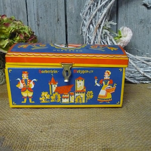 Könnte beinhalten: Eine blaue Vintage-Metallbox mit Klappdeckel, mit Illustrationen von Personen, einem Schloss und dem Wort "Nürnberg". Die Box hat rote und gelbe Akzente und einen Metallverschluss.