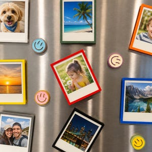 May include: A collection of rectangular magnetic photo frames in various colors including white, green, orange, yellow, red, blue, gray, and black, each holding a different photograph, accompanied by several small round smiley face magnets.