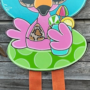 May include: A whimsical flamingo-shaped wooden door hanger. The flamingo wears sunglasses, a sun hat, and a leopard print bikini top. It holds a cocktail with a tiny umbrella and a lemon slice. The design features bright pink, blue, green, and orange colors.