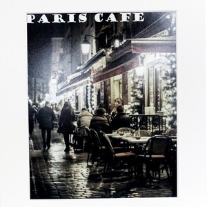 May include: A black and white photo of a Parisian cafe at night with people sitting at tables outside. The cafe is decorated with string lights and the sign above the entrance reads "PARIS CAFE".