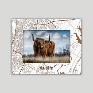May include: A framed photograph of a brown Highland cow standing in a field with a cloudy sky in the background. The frame is made of wood and has a map of Austin, Texas printed on it. The word "Austin" is printed in brown letters at the bottom of the frame.