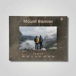 May include: A brown wooden frame with a topographic map design featuring Mount Rainier. The frame includes a photo of two people standing on a mountaintop with a cloudy background. The text "Mount Rainier", "14,411 ft", "46° 51' 11" N 121° 45' 38" W" is printed on the frame.