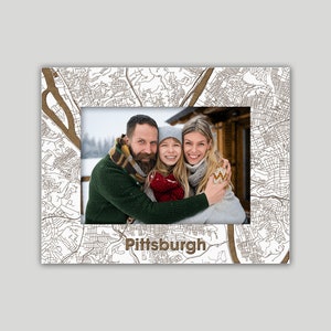 May include: A white photo frame with a brown outline of a city map. The map is of Pittsburgh, Pennsylvania. The frame has a photo of a family of three in the center. The family is smiling and hugging each other. The photo is taken in a snowy setting.