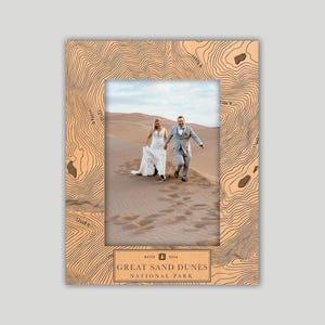 May include: A wooden picture frame with a topographic map design. The frame surrounds a photo of a couple running on a sand dune. The text "GREAT SAND DUNES NATIONAL PARK" is at the bottom.