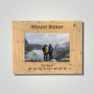 May include: A wooden photo frame with a topographic map design featuring Mount Baker, Washington. The frame includes the mountain's elevation, 10,786 feet, and coordinates: 48° 46' 36" N 121° 48' 52" W. The frame has a photo of two people standing on a mountaintop with fog in the background.