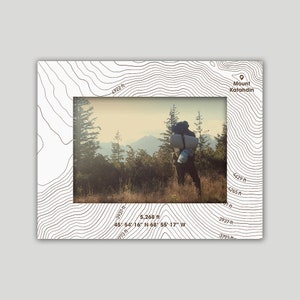 May include: A black and white topographic map with brown text and a photo of a person hiking in a forest. The text on the map reads "Mount Katahdin" and "5,268 ft 45° 54' 16" N 68° 55' 17" W".