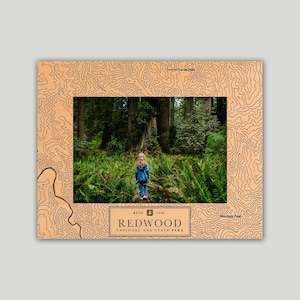 May include: A rectangular wooden picture frame featuring a laser-engraved topographic map pattern. The center displays a photograph of a person in a blue jacket standing in a forest. Text reads ESTD 1968 REDWOOD NATIONAL AND STATE PARK.