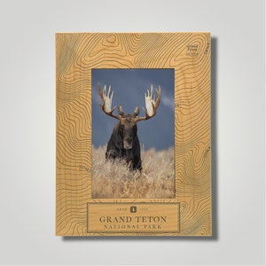 May include: A wooden photo frame with a topographic map design featuring the Grand Teton National Park. The frame has a photo of a moose with large antlers in the center. The text "ESTD 1929 GRAND TETON NATIONAL PARK" is printed on the bottom of the frame.