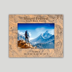 May include: A wooden frame with a topographic map design surrounds a photograph of Mount Everest. The photo shows a person standing on a rocky outcrop, with the mountain in the background. Text on the frame reads "Mount Everest South Base Camp" and "17,598 ft".
